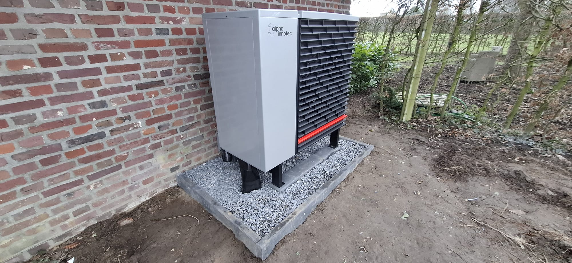 Außeneinheit einer Wärmepumpe der Marke alpha innotec auf Schotterfundament vor einer Backsteinwand installiert.